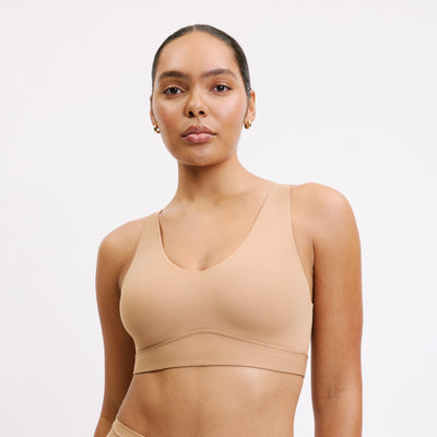 On a white studio backdrop, Amber wears the Bliss Bralette in Tan, shown in a centered front view. The wireless design, scoop neckline, and lightweight microfiber fabric are highlighted.