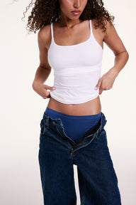 Against a light studio backdrop, Courtney wears the Bliss Cami in White with blue underwear and unbuttoned jeans. The scoop neckline, thin adjustable straps, and ultra-soft microfiber fabric create a smooth, stretchy fit. sizing: Courtney wears size Small