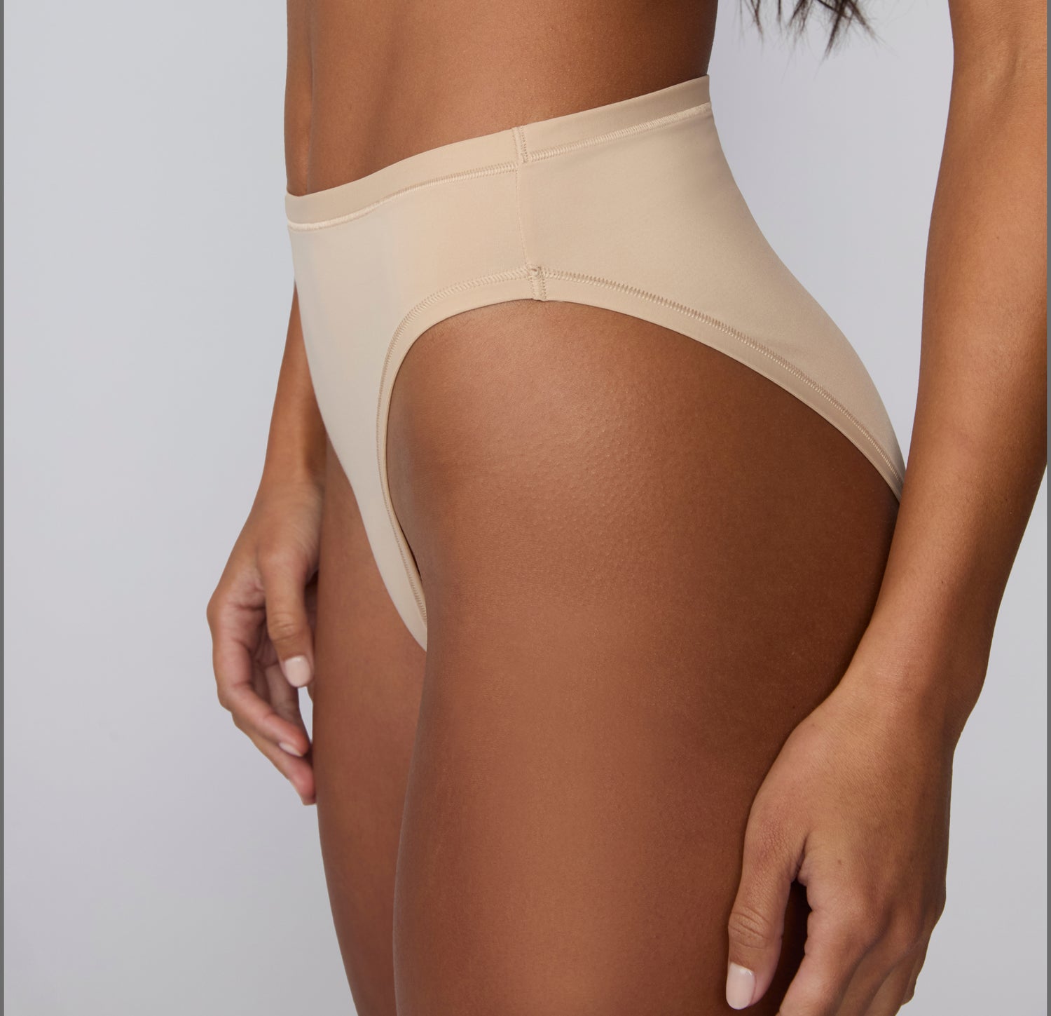 On a light gray studio backdrop, Amber wears the Bliss French Cut Brief in Beige, shown from a side angle to highlight the smooth waistband and high-cut leg opening.