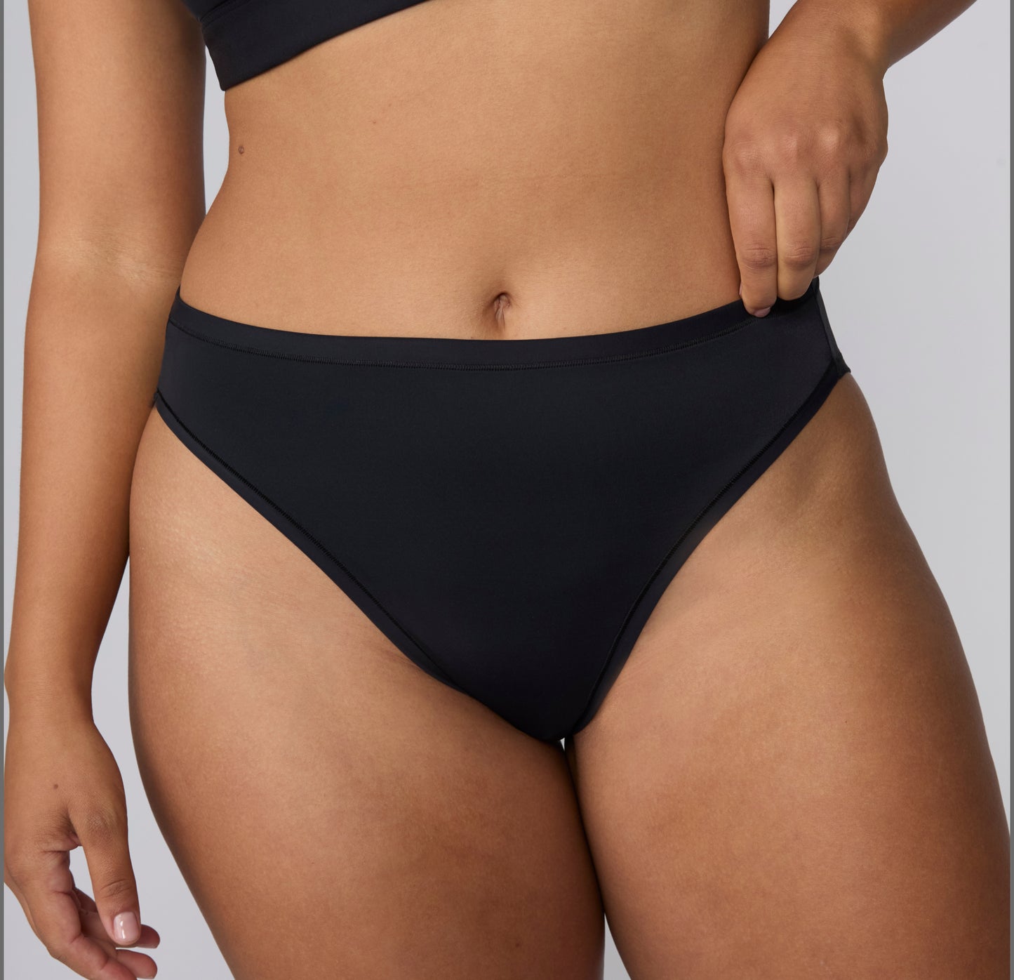 On a light gray studio backdrop, a close-up of India wearing the Bliss French Cut Brief in Black shows the smooth front panel, mid-rise waistband, and high-cut leg opening.