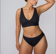 On a light gray studio backdrop, India wears the Bliss French Cut Brief in Black with the matching bra, shown in a three-quarter view to highlight the soft microfiber fabric and clean edges.