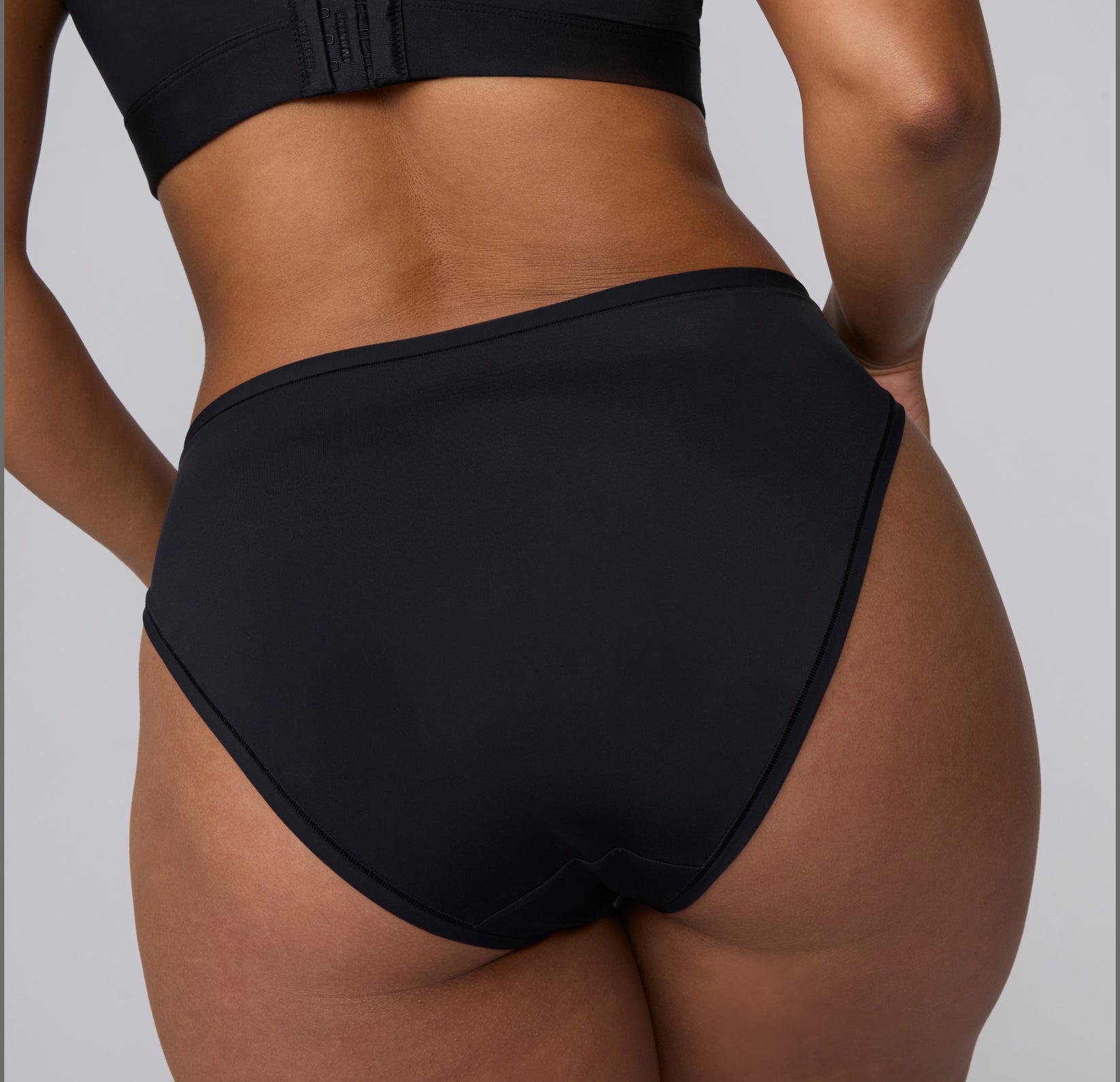 On a light gray studio backdrop, a back view of India wearing the Bliss French Cut Brief in Black shows the smooth rear coverage and clean finish under the fabric.
