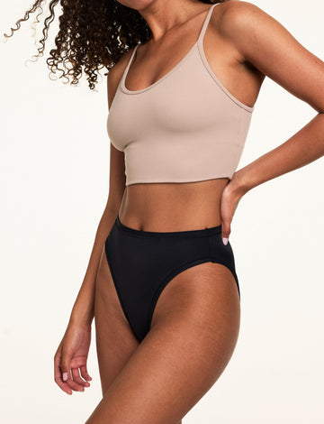 On a light studio backdrop, Courtney wears the Bliss French Cut in Black with a beige bralette. The underwear features a high-rise silhouette, high-cut leg openings, and smooth microfiber fabric.