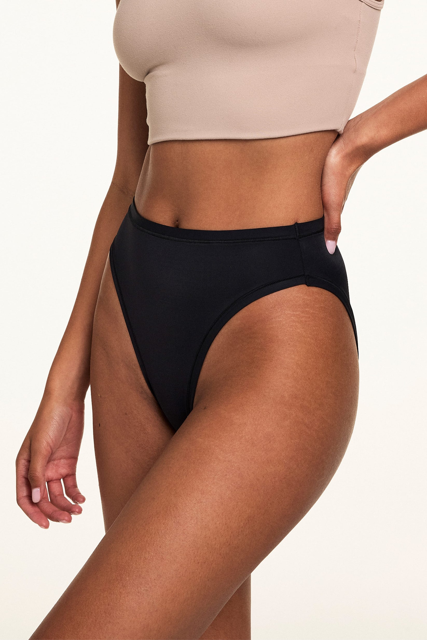 Close-up side view on a light studio backdrop, Courtney wears the Bliss French Cut in Black with a beige bralette. The underwear features a high-rise fit, high-cut leg openings, and smooth microfiber fabric.