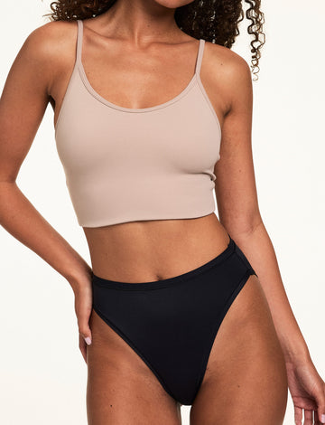 On a light studio backdrop, Courtney wears the Bliss French Cut in Black with a beige bralette. The underwear features a high-rise silhouette, smooth microfiber fabric, and high-cut leg openings. sizing: Courtney wears size Small
