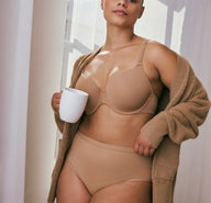 In a softly lit room, a model wears the Bliss Highwaist Brief in Tan, paired with a matching lightly lined bra. The high-rise waistband sits smooth against the body, showing the soft, no-show fabric. The model holds a white mug and wears a cozy cardigan draped over her shoulders.