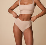 Ally wearing the Bliss Highwaist Brief in Beige, standing against a soft beige background. The brief features a high-rise fit with smooth, minimal edges for all-day comfort and a flattering silhouette. sizing: Ally wears size Small