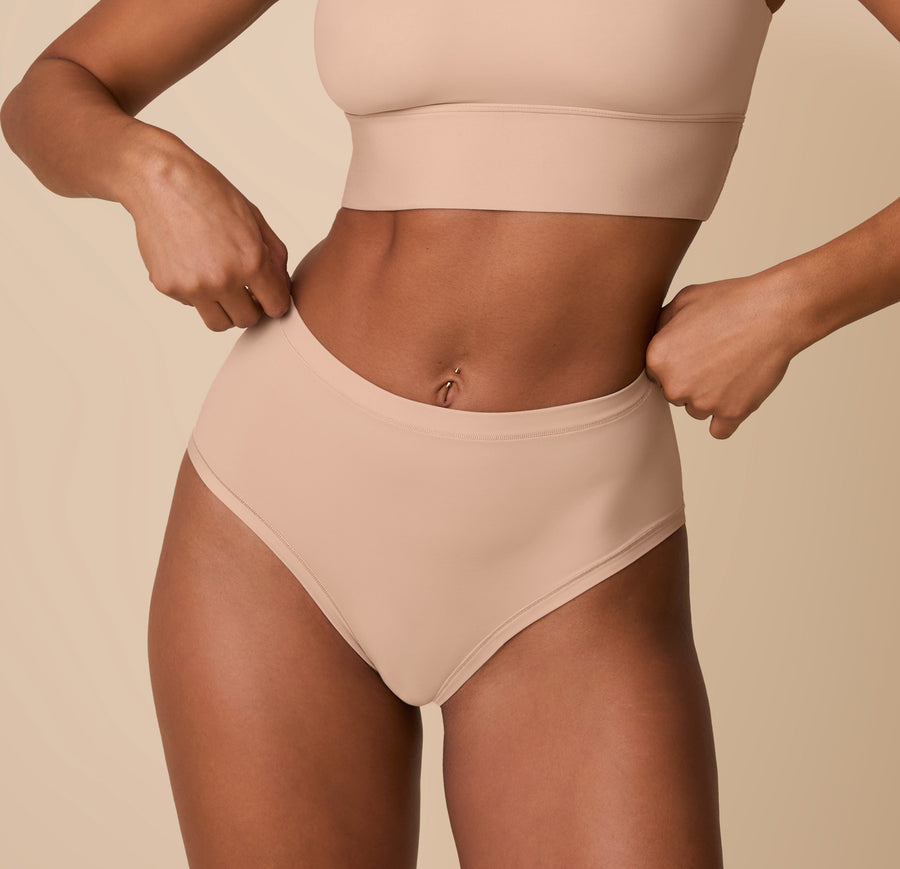 Ally wearing the Bliss Highwaist Brief in Beige, standing against a soft beige background. The brief features a high-rise fit with smooth, minimal edges for all-day comfort and a flattering silhouette. sizing: Ally wears size Small