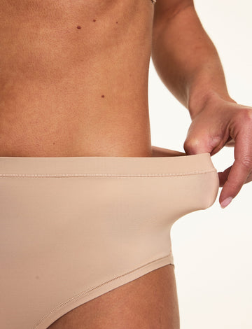 Close-up on a light studio backdrop, Gianna stretches the waistband of the Bliss Highwaist in Beige to show the flexible microfiber fabric and high-rise fit.