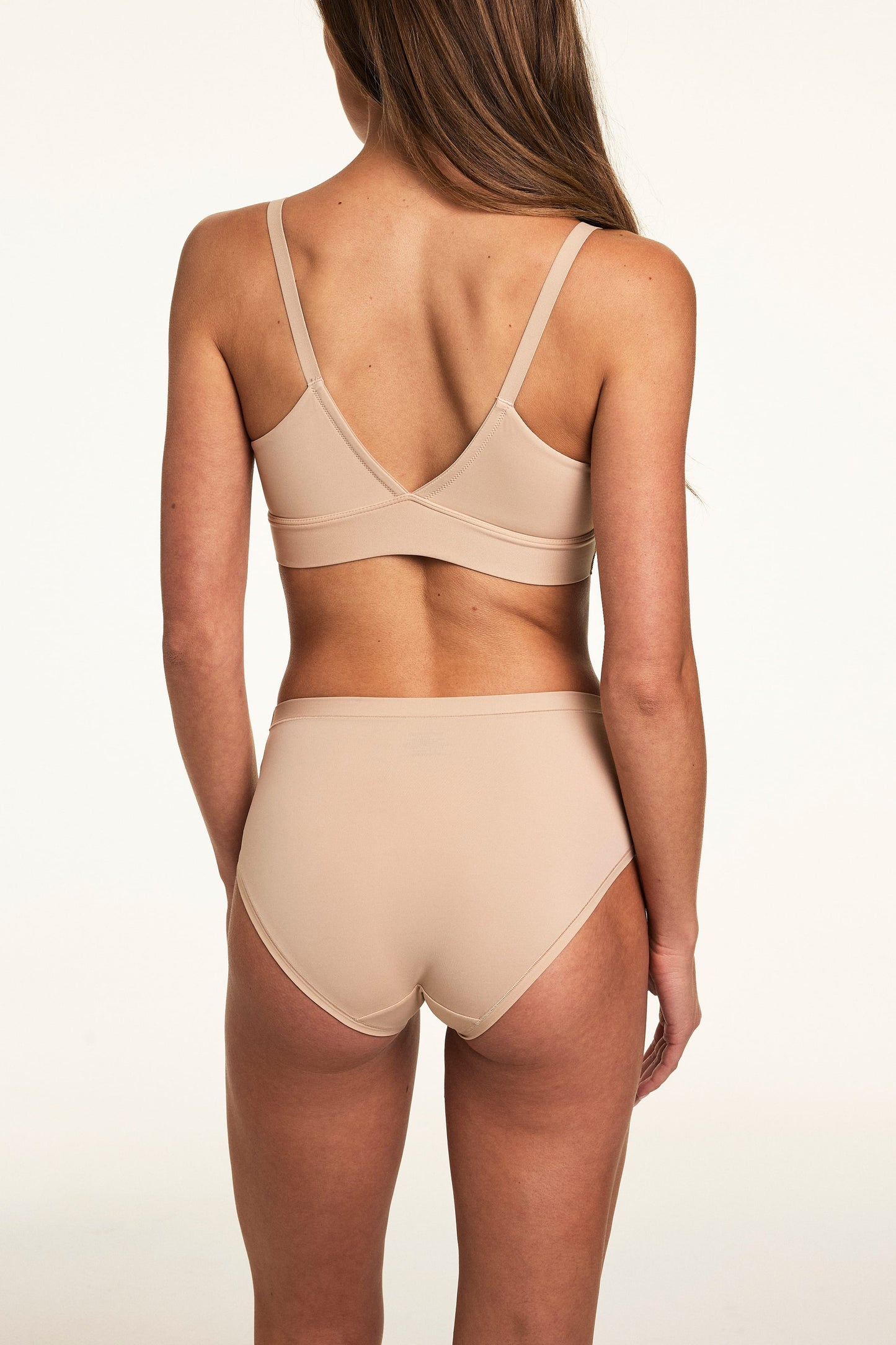 Back view on a light studio backdrop, Gianna wears the Bliss Highwaist in Beige with the Bliss Triangle Bralette in Beige. The underwear features full back coverage, a smooth waistband, and soft microfiber fabric.