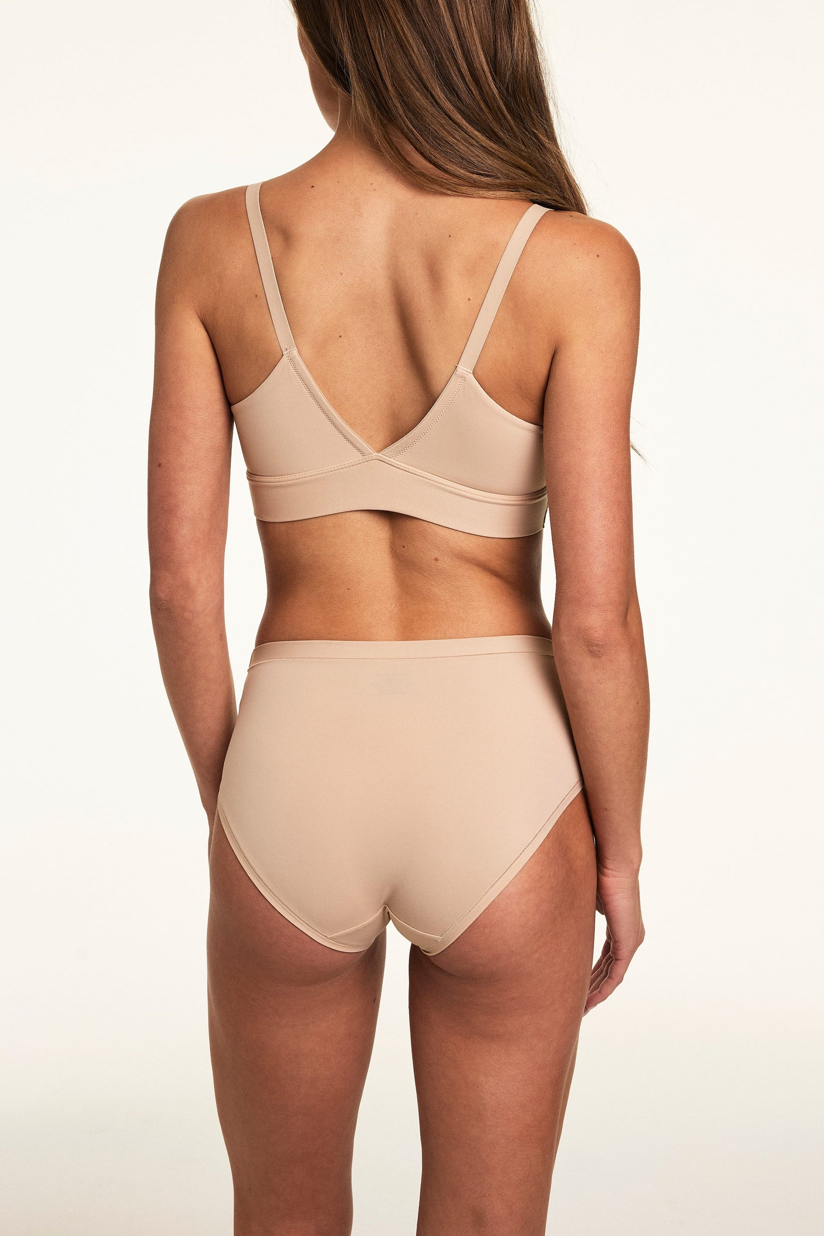 Back view on a light studio backdrop, Gianna wears the Bliss Highwaist in Beige with the Bliss Triangle Bralette in Beige. The underwear features full back coverage, a smooth waistband, and soft microfiber fabric.