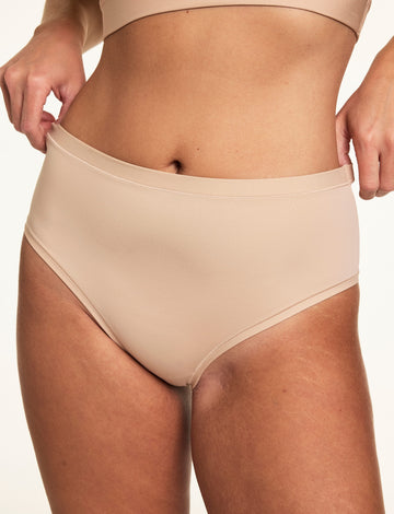 Close-up on a light studio backdrop, Sonnie stretches the waistband of the Bliss Highwaist in Beige to show the flexible microfiber fabric and high-rise fit.