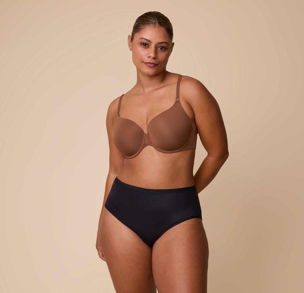 India wearing the Bliss Highwaist Brief in Black paired with the Base T-Shirt Bra in Brown, standing against a neutral backdrop with a confident, relaxed expression.