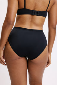 On a light grey studio backdrop, Amber wears the Bliss Highwaist in Black, back view. The high-rise design provides full back coverage with smooth, elastic-free edges and soft microfiber fabric that lays flat against the skin.