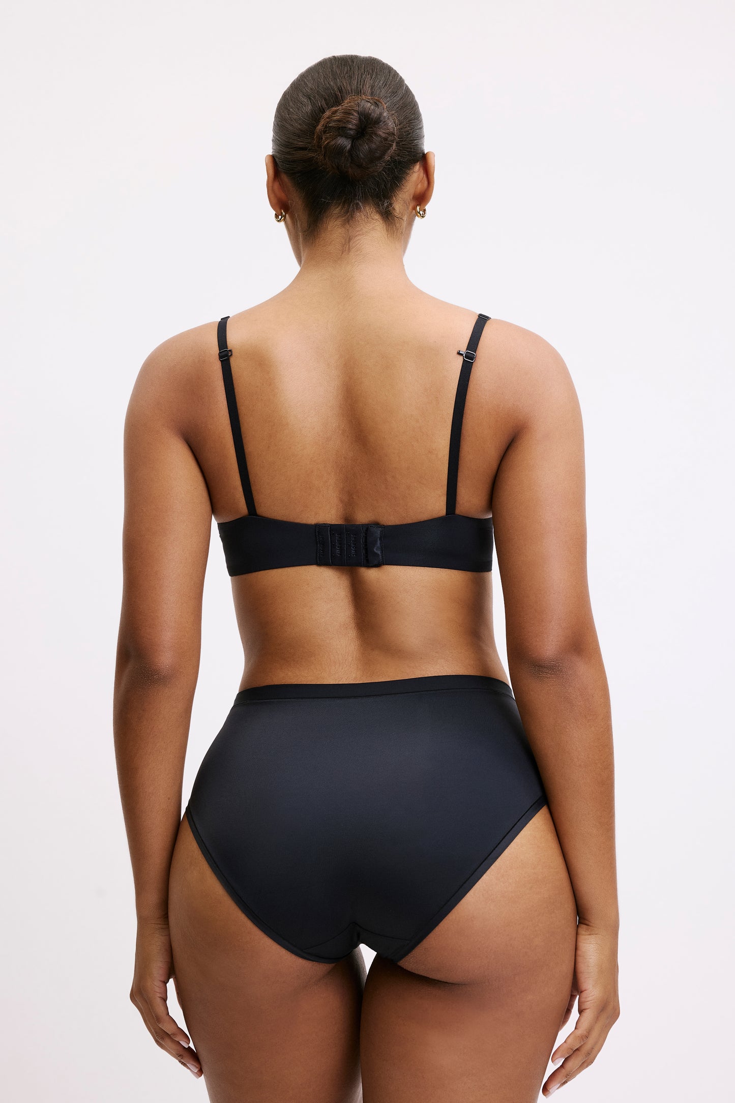 On a light grey studio backdrop, back view of Amber wearing the Bliss Highwaist in Black paired with a matching black bra. The high-waisted silhouette offers full coverage with smoothing microfiber fabric designed to disappear under clothing.