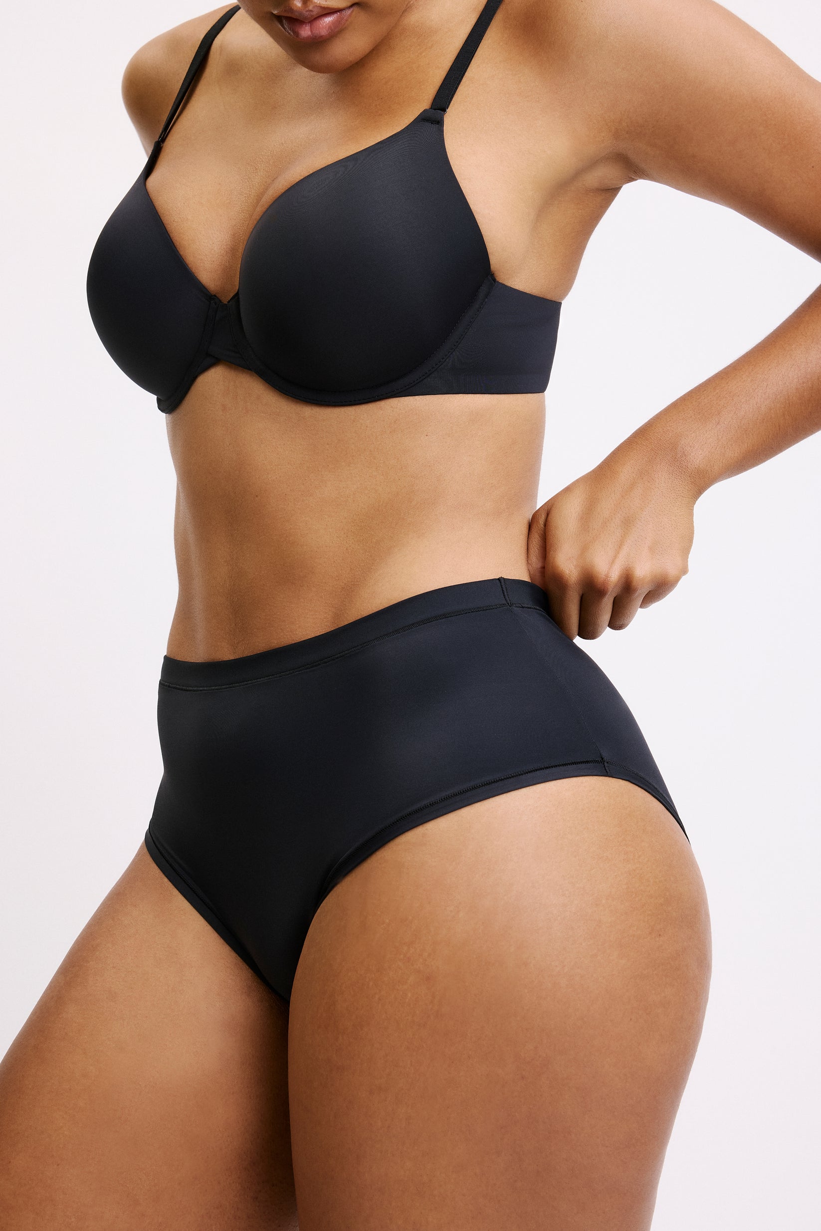 On a light grey studio backdrop, Amber wears the Bliss Highwaist in Black, angled front view. The high-rise waistband sits smoothly at the natural waist, showcasing the ultra-soft microfiber fabric and full front coverage designed to disappear under clothing.