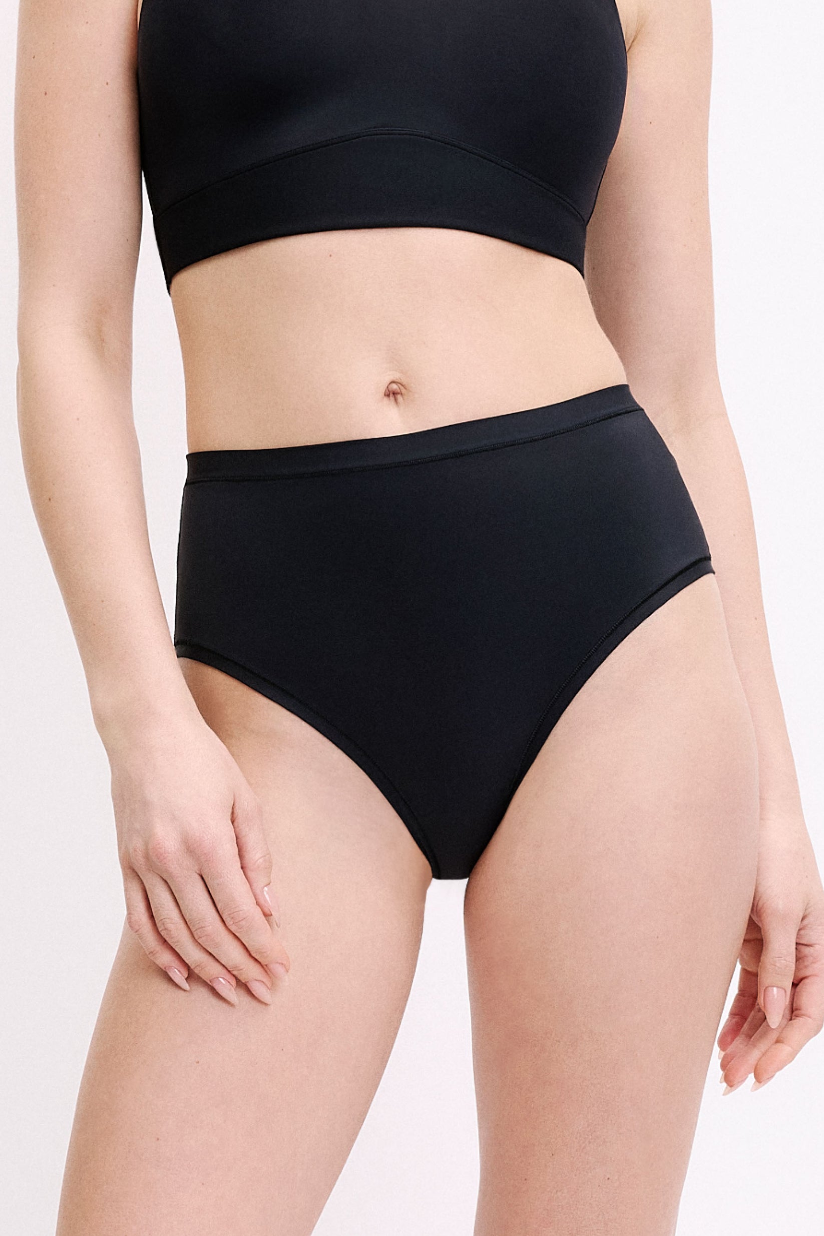 On a white studio backdrop, a front close-up shows Megan wearing the Bliss Highwaist Brief in Black. The smooth waistband and full front coverage are visible, designed to sit comfortably at the waist. sizing: Megan wears size Small