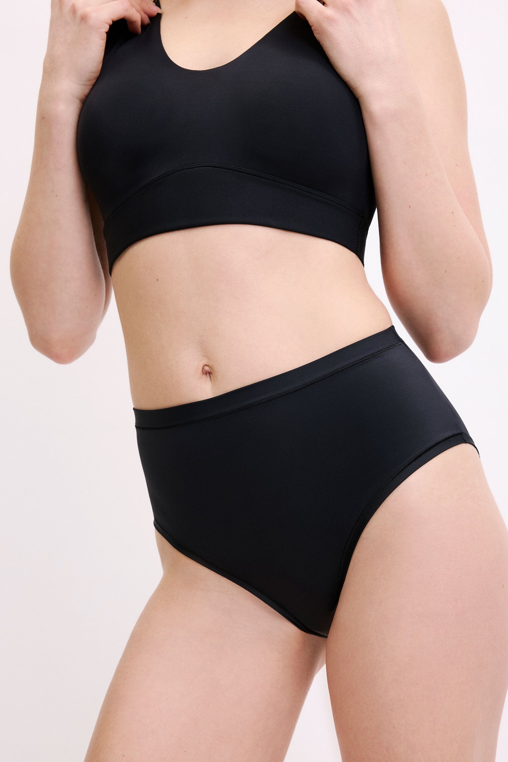 On a white studio backdrop, a three-quarter front close-up shows Megan wearing the Bliss Highwaist Brief in Black. The smooth waistband and clean leg openings are visible, designed for everyday comfort.