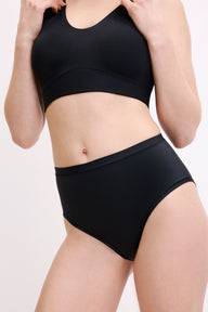 On a white studio backdrop, a three-quarter front close-up shows Megan wearing the Bliss Highwaist Brief in Black. The smooth waistband and clean leg openings are visible, designed for everyday comfort.
