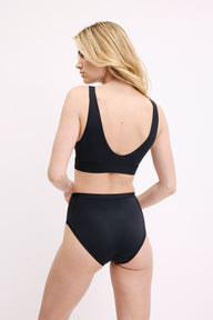On a white studio backdrop, Megan is shown from a three-quarter back view wearing the Bliss Highwaist Brief in Black, paired with the Bliss Bralette. The full back coverage and soft microfiber fabric are visible.