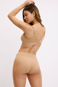 On a white studio backdrop, Darion is shown from the back wearing the Bliss Triangle Bralette and Bliss Highwaist Brief in Tan, showing the bralette’s supportive back band and the brief’s full back coverage with a smooth, elastic-free finish.