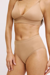 On a white studio backdrop, Darion is shown in a close-up front view wearing the Bliss Triangle Bralette and Bliss Highwaist Brief in Tan, highlighting the deep V neckline, smooth underband, and the brief’s high-rise waist in soft microfiber fabric.