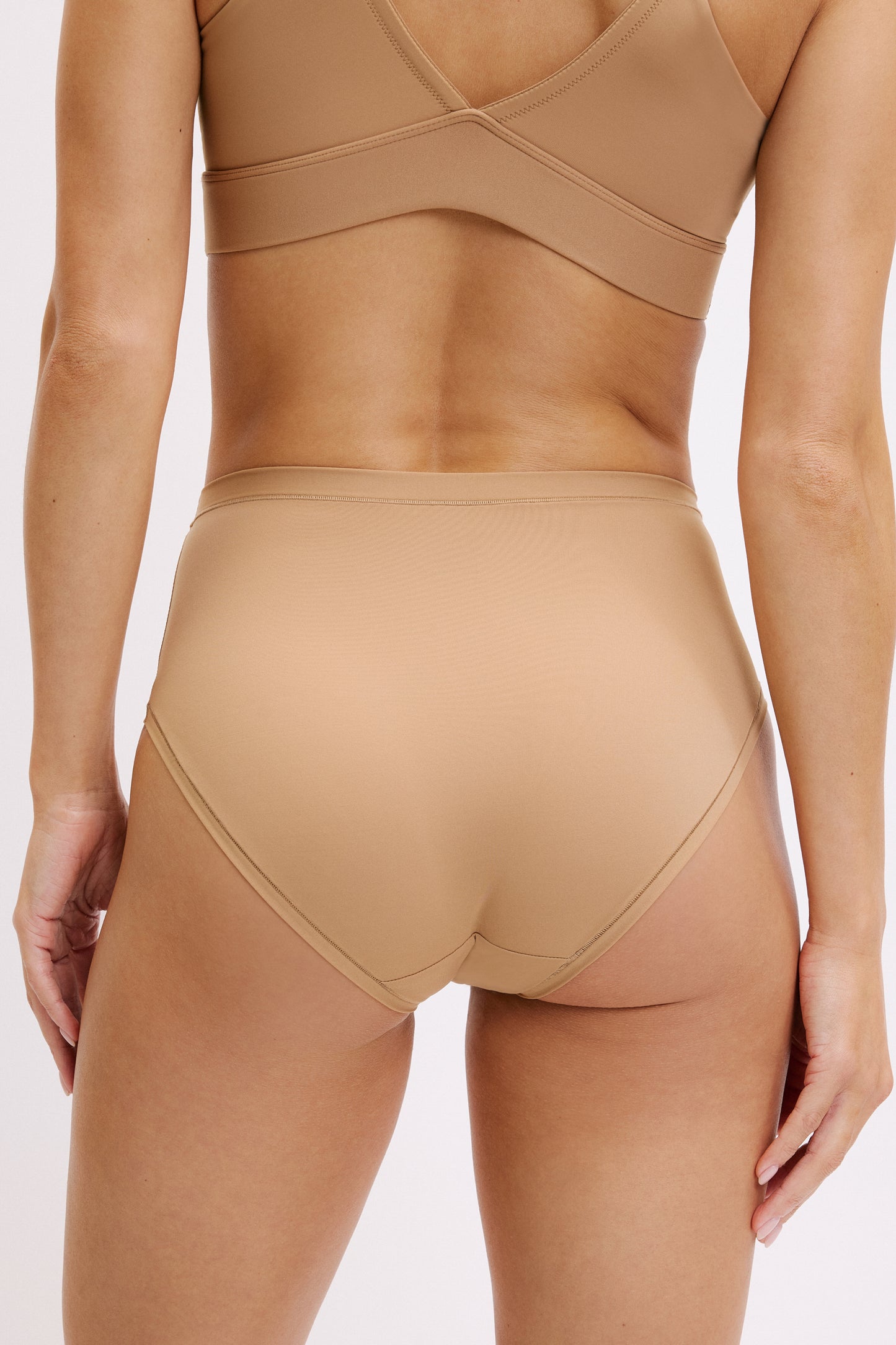 On a white studio backdrop, Darion is shown in a close-up back view wearing the Bliss Triangle Bralette and Bliss Highwaist Brief in Tan, showing the bralette’s supportive back band and the brief’s full back coverage with a smooth, elastic-free finish.