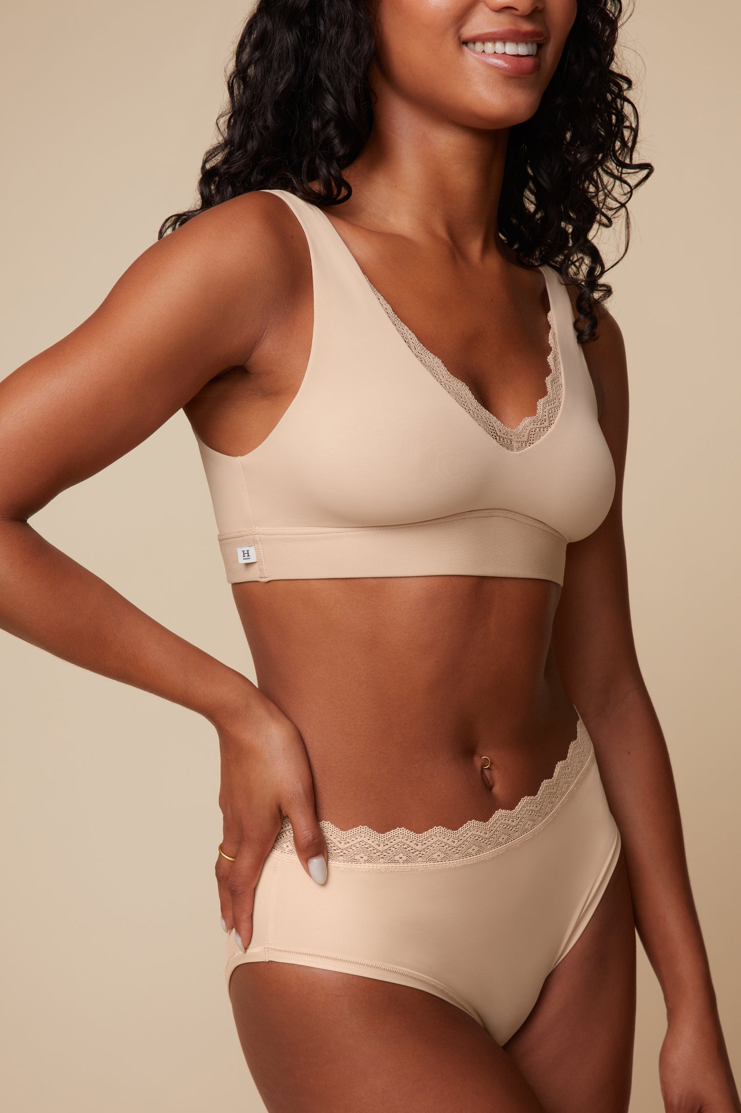 On a warm beige studio backdrop, Ally wears the Bliss Lace Bralette in Beige, front view. The V-neck silhouette is finished with soft, stretchy lace trim and smoothing microfiber fabric for wireless lift and all-day ease. sizing: Ally wears size Small