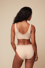 On a warm beige studio backdrop, back view of Ally wearing the Bliss Lace Bralette in Beige with matching underwear. The bralette features a low V-back with soft lace trim and a smooth wireless band designed to lay flat against the body.