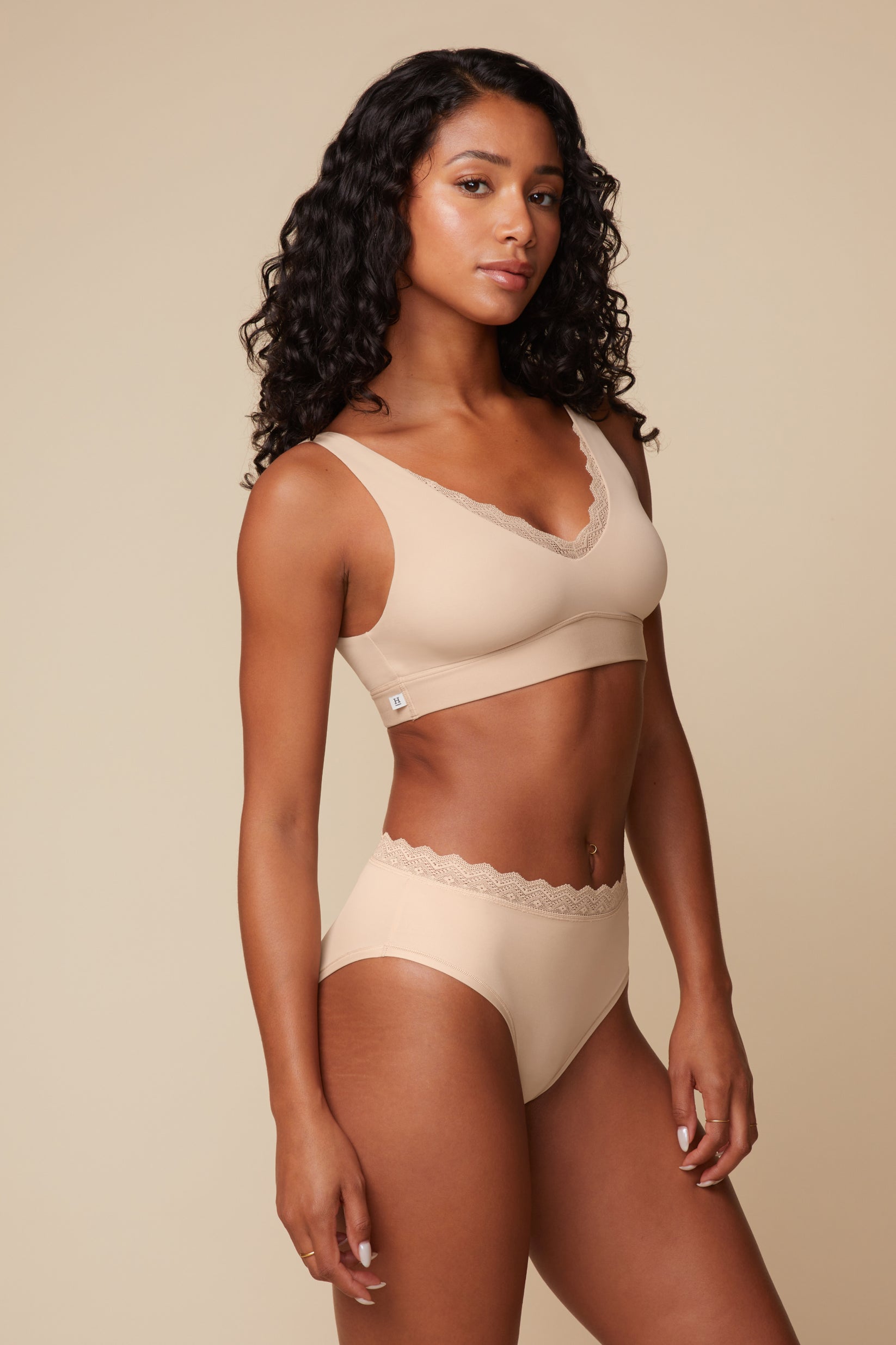 On a warm beige studio backdrop, Ally wears the Bliss Lace Bralette in Beige with matching underwear, angled front view. The ultra-soft fabric and scalloped lace trim create a smooth silhouette with a comfortable wireless fit.