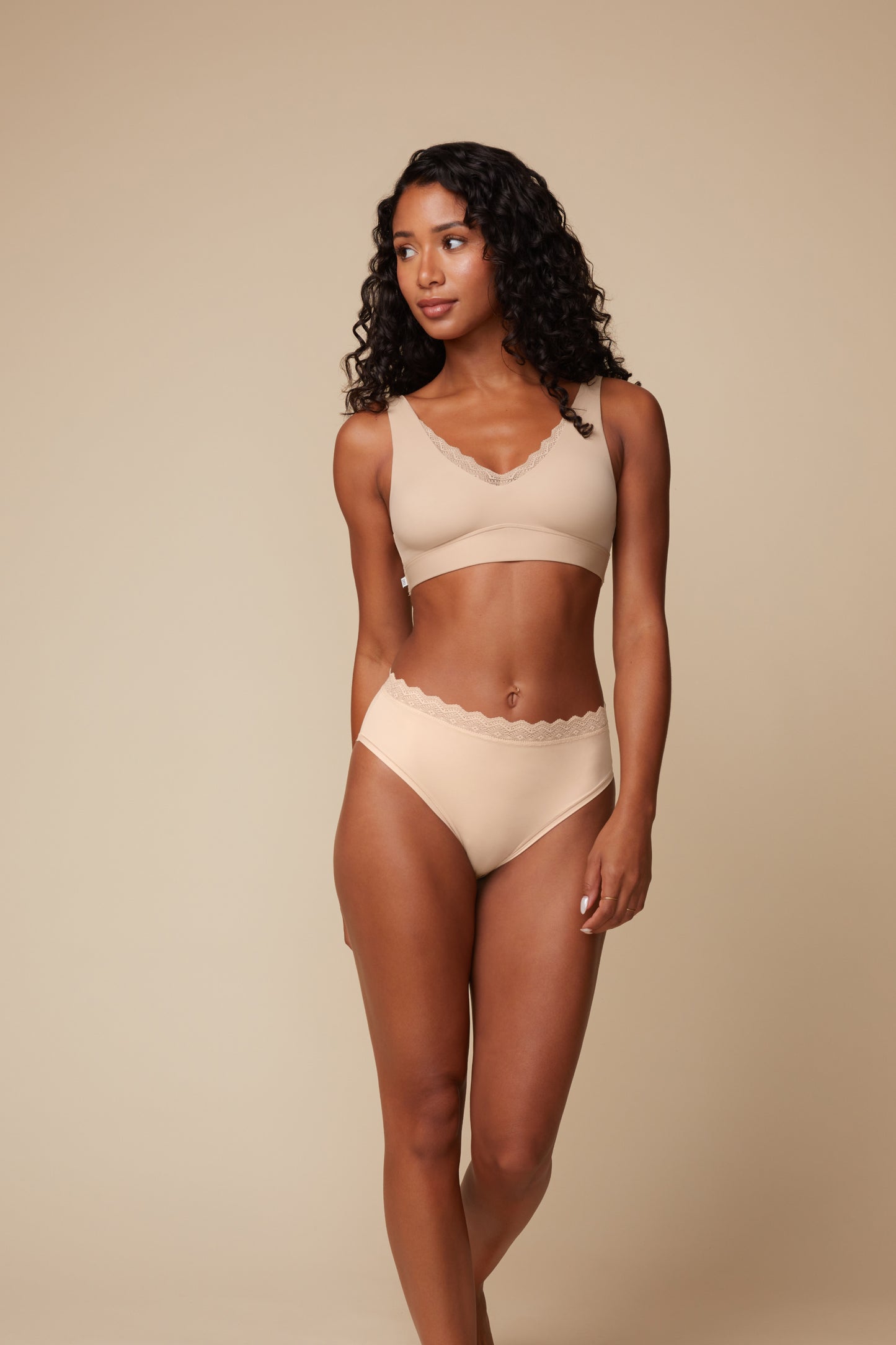 On a warm beige studio backdrop, Ally wears the Bliss Lace Bralette in Beige with matching underwear, full-length front view. The wireless bralette features a soft V-neckline trimmed in delicate lace and a smooth underband designed for everyday comfort.