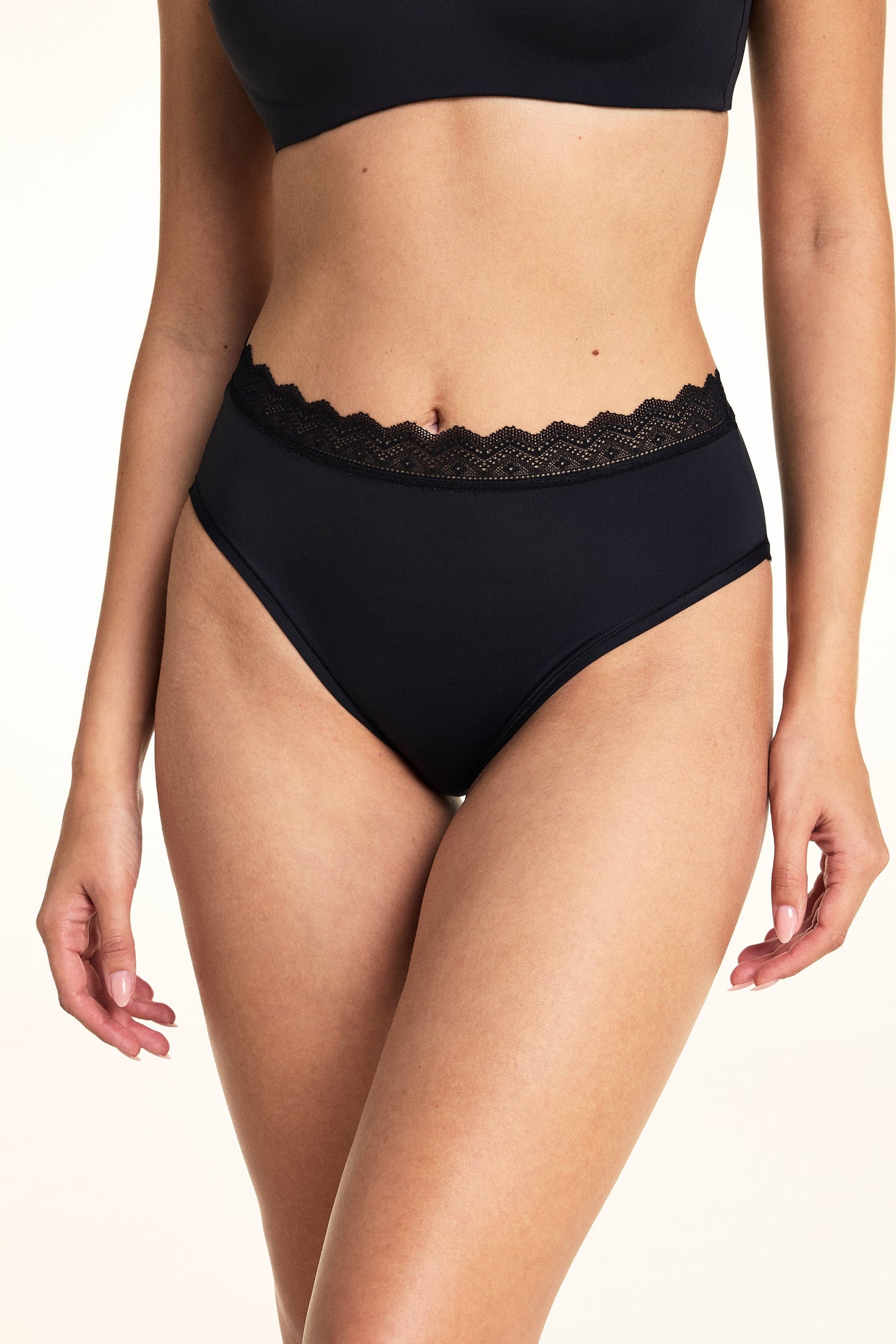 In a studio setting, Laura wears the Bliss Lace Highwaist Brief in Black, highlighting the high-rise silhouette, smooth microfiber fabric, and scalloped lace trim along the waistband. sizing: Laura wears size Small