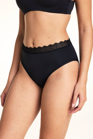 In a studio setting, Laura wears the Bliss Lace Highwaist Brief in Black, highlighting the high-rise silhouette, smooth microfiber fabric, and scalloped lace trim along the waistband from an angled front view.