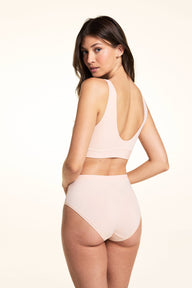 In a studio setting, Laura wears the Bliss Lace Highwaist Brief in Rose from the back, featuring a high-rise silhouette, smooth microfiber fabric, and full back coverage with a clean finish.