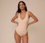 Marcela facing forward with hands on hips in the Bliss Plunge Bodysuit in Beige. The image emphasizes the sleek, supportive fit and neutral tone of the bodysuit.