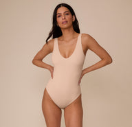 Marcela facing forward with hands on hips in the Bliss Plunge Bodysuit in Beige. The image emphasizes the sleek, supportive fit and neutral tone of the bodysuit.