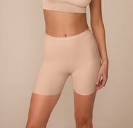 Front view of Marcela wearing the Bliss Under Short in Beige. The high-rise short extends to mid-thigh, showcasing its smooth, tag-free design and soft microfiber fabric. sizing: Marcela wears size Small