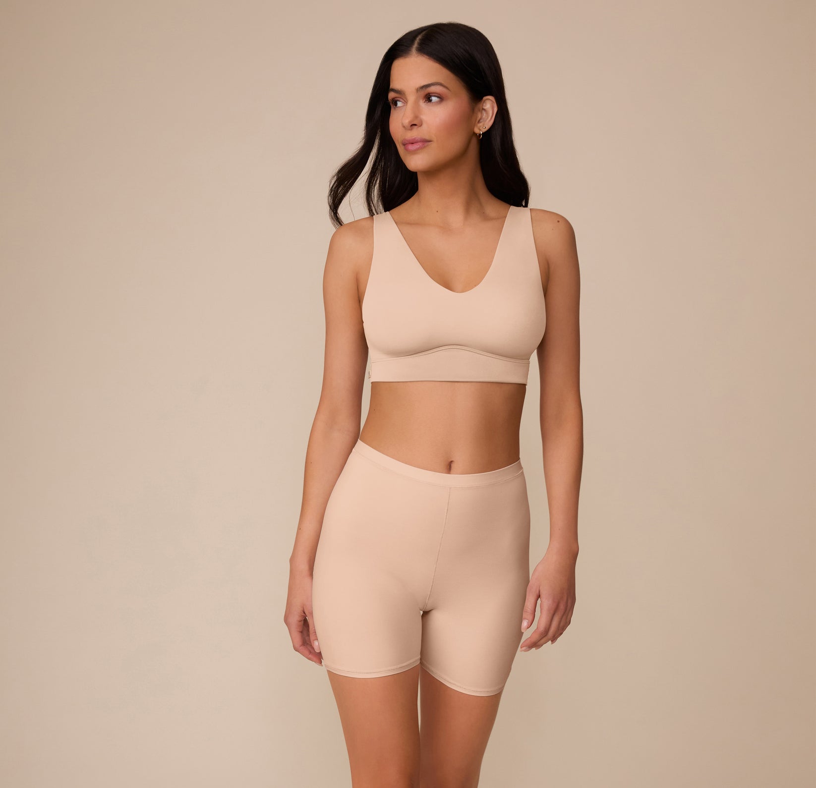 Marcela standing with a soft expression, wearing the Bliss Under Short and matching Bliss Bralette in Beige. The full set highlights the smooth, supportive fit and neutral tone.
