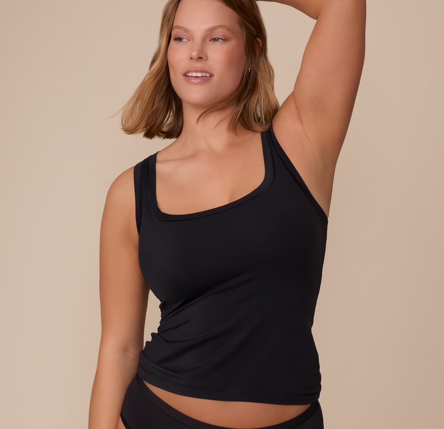 Front view of Sonnie wearing the Bliss Tank in Black, one arm raised. The tank features a wide scoop neckline and a close, comfortable fit. sizing: Sonnie wears size Medium