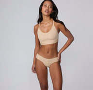 On a light gray studio backdrop, Amber wears the Bliss Low Rise Bikini in Beige with the matching bralette, showing the full set with a soft, supportive waistband and minimal, streamlined design.