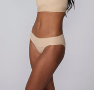 On a light gray studio backdrop, Amber wears the Bliss Low Rise Bikini in Beige, shown at an angled side view to show the low-rise waistband and seamless-looking edges under clothing.
