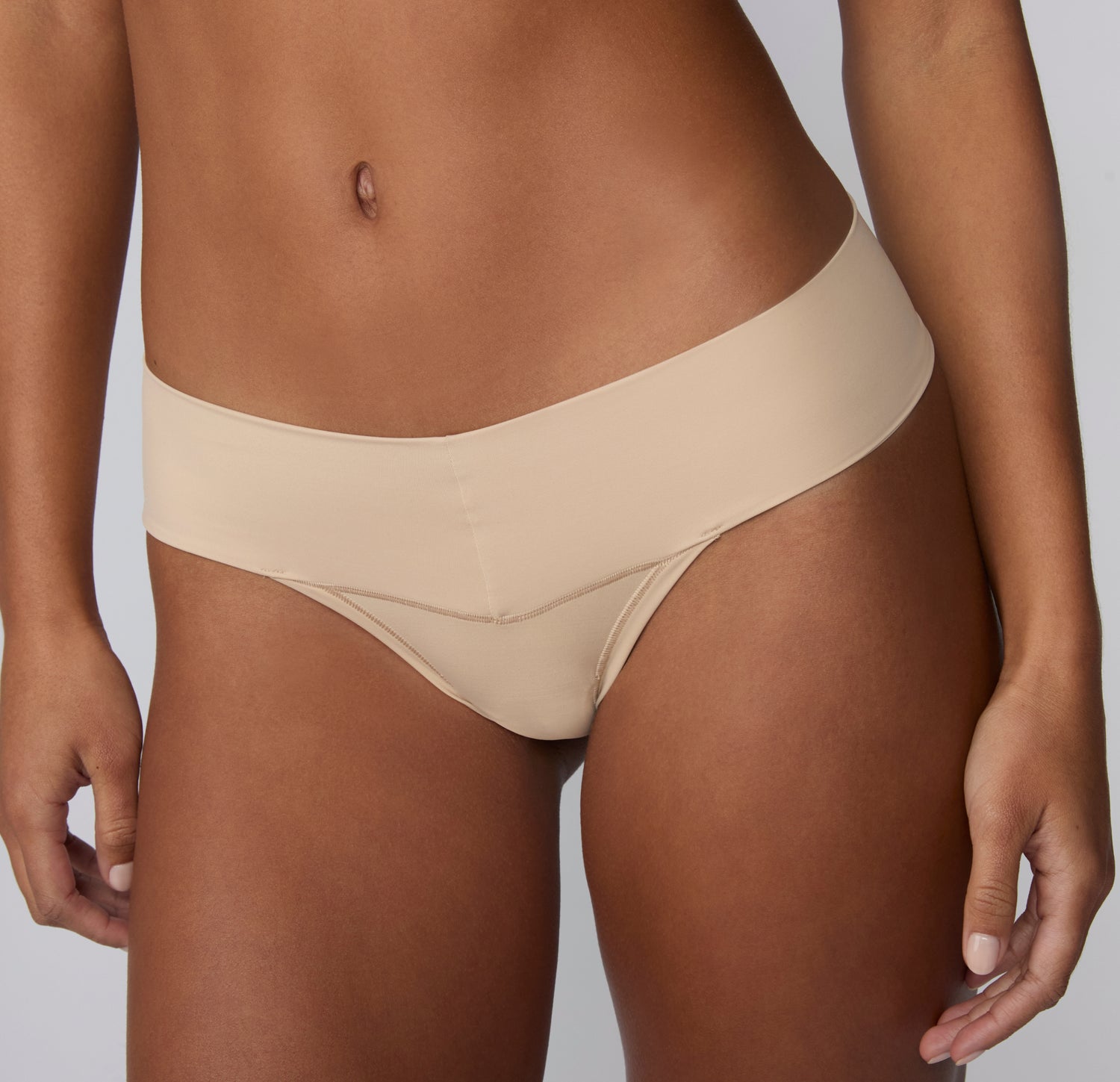 On a light gray studio backdrop, a front close-up of Amber wearing the Bliss Low Rise Thong in Beige shows the smooth microfiber fabric and low-rise waistband.