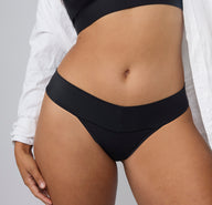 On a light gray studio backdrop, a front close-up of India wearing the Bliss Low Rise Thong in Black shows the smooth microfiber fabric and low-rise waistband.