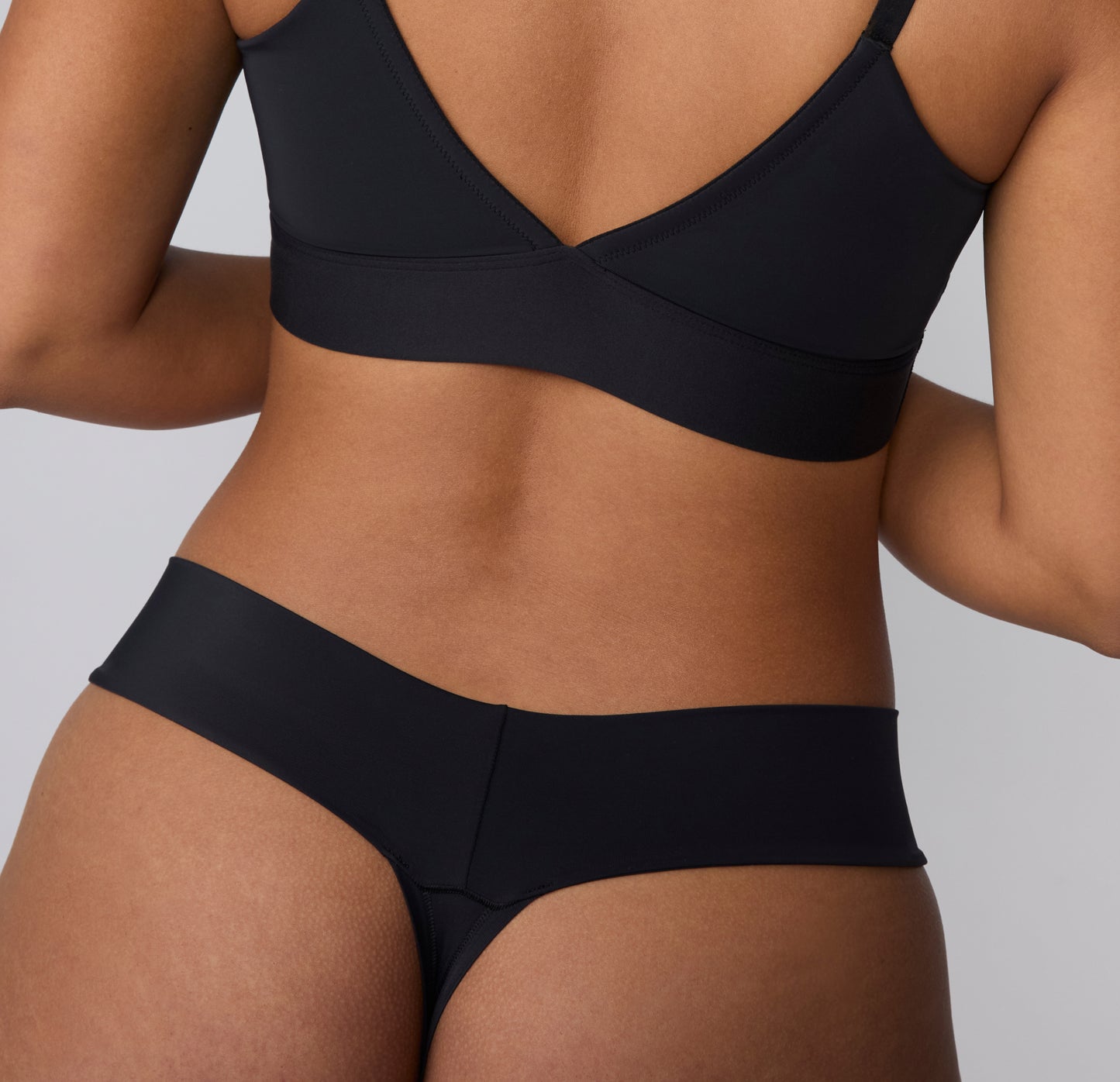 On a light gray studio backdrop, a back view of India wearing the Bliss Low Rise Thong in Black highlights the smooth fabric and minimal back coverage.