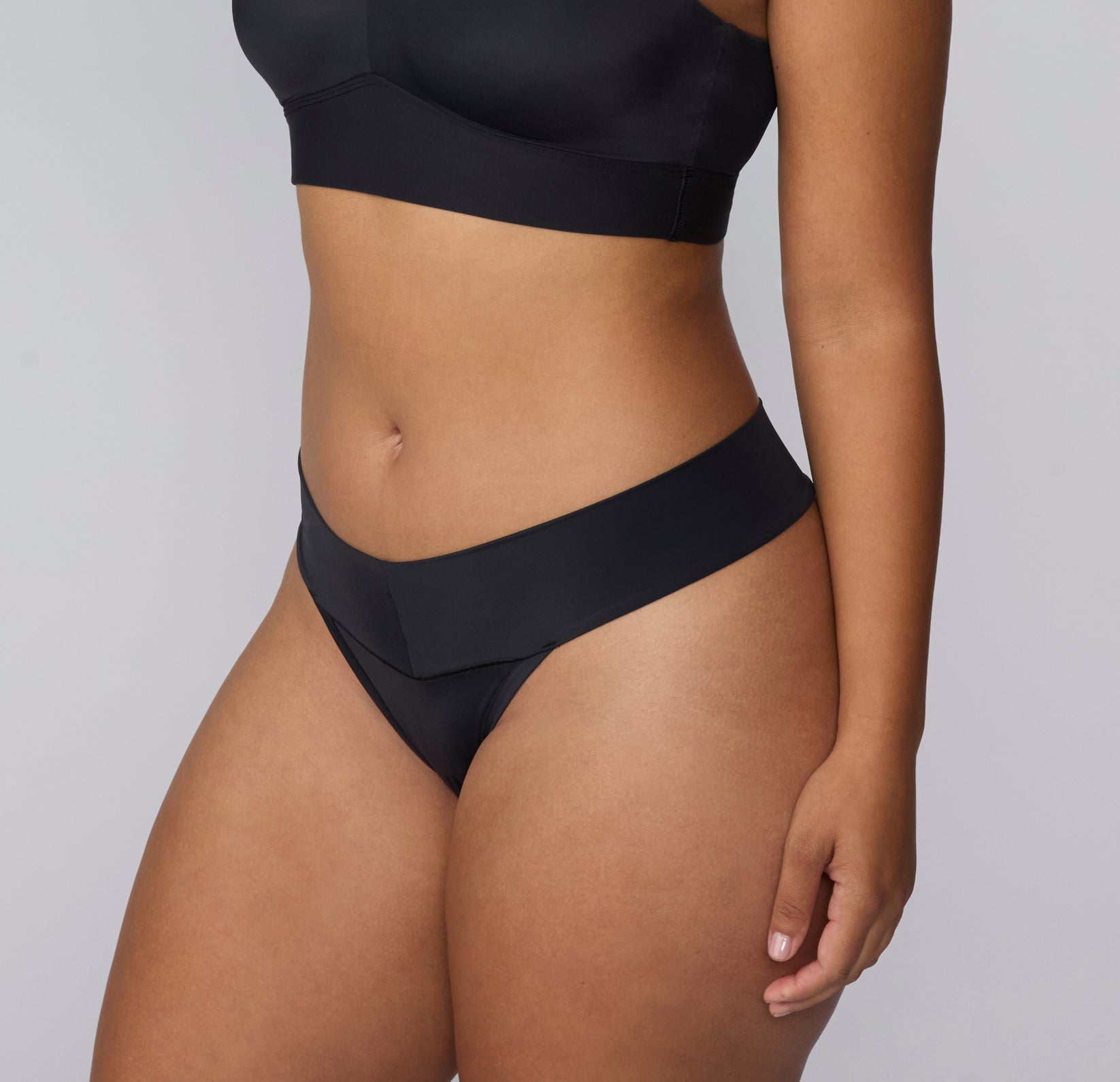 On a light gray studio backdrop, a three-quarter front view of India wearing the Bliss Low Rise Thong in Black shows the low-rise silhouette and clean waistband finish.