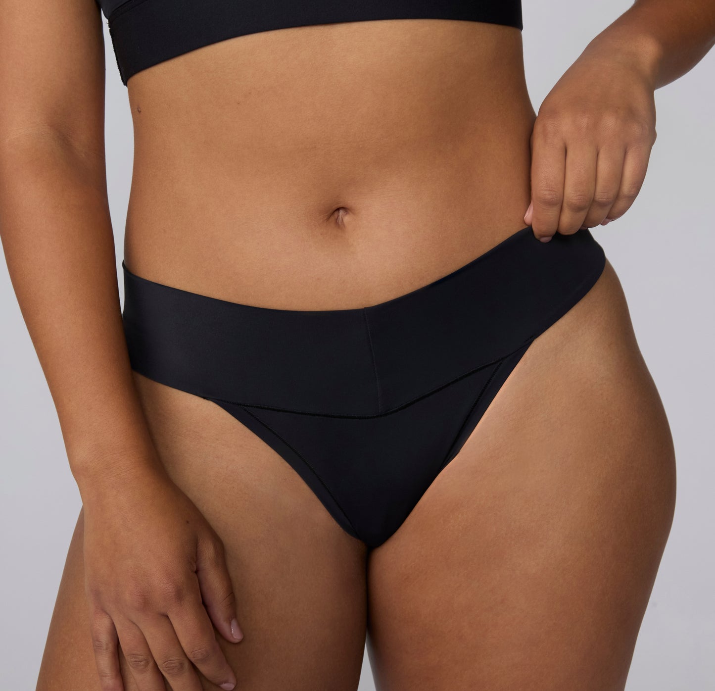 On a light gray studio backdrop, a front close-up of India wearing the Bliss Low Rise Thong in Black highlights the soft microfiber fabric and smooth edges.