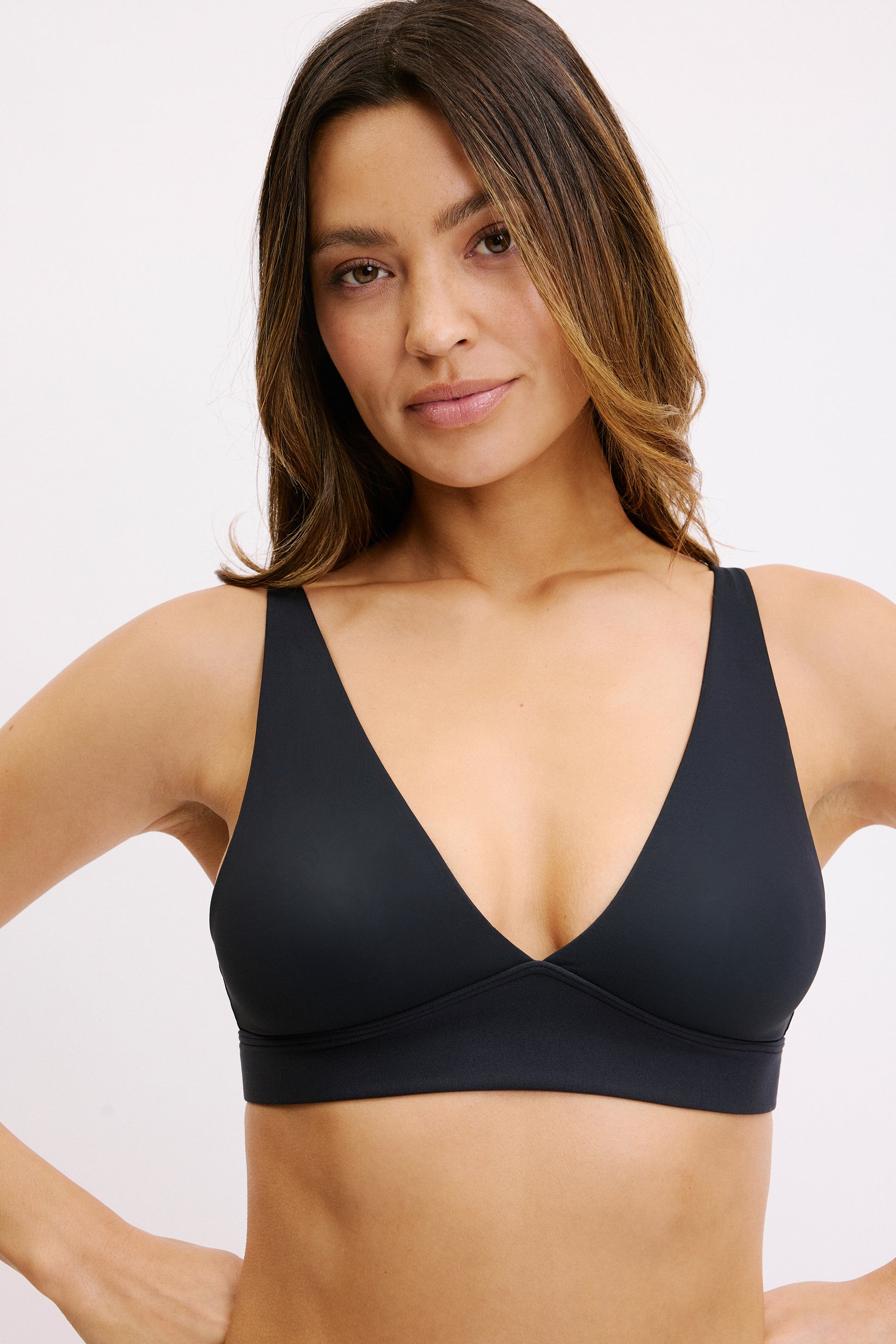 On a white studio backdrop, Darion wears the Bliss Plunge Adjustable Bralette in Black, shown in a close-up front view. The deep plunge neckline and smooth microfiber fabric are visible, with wide straps and a clean underband. sizing: Darion wears size Small