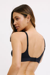 On a white studio backdrop, Darion is shown from a three-quarter back angle wearing the Bliss Plunge Adjustable Bralette in Black, showing the adjustable shoulder straps, supportive back band, and clean, wire-free construction.