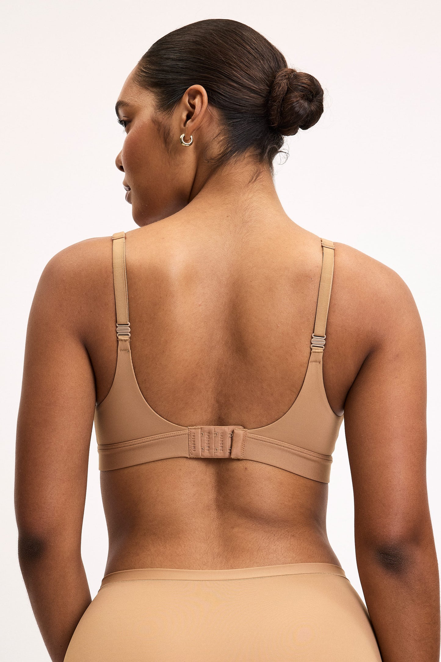 Back view of Amber wearing the Bliss Plunge Adjustable Bra in Tan with matching Tan underwear, highlighting the adjustable straps, hook-and-eye closure, and smooth scoop back. 