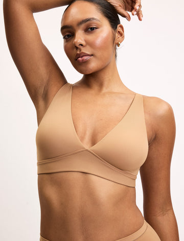 Amber wears the Bliss Plunge Adjustable Bra in Tan with matching Tan underwear, raising one arm to highlight the smooth fabric, deep V-neckline, and supportive underband. sizing: Amber wears size Medium